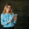 Smiling businesswoman with tablet looking at camera Happy beautiful young business woman standing with tablet, widely smiling. Dressed in elegant blouse, in eyeglasses.